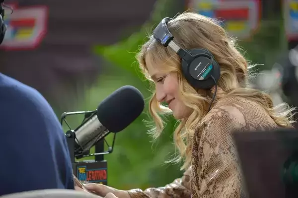 Woman with blond hair in front of a microphone wearing headphones in front of an outdoor set background.