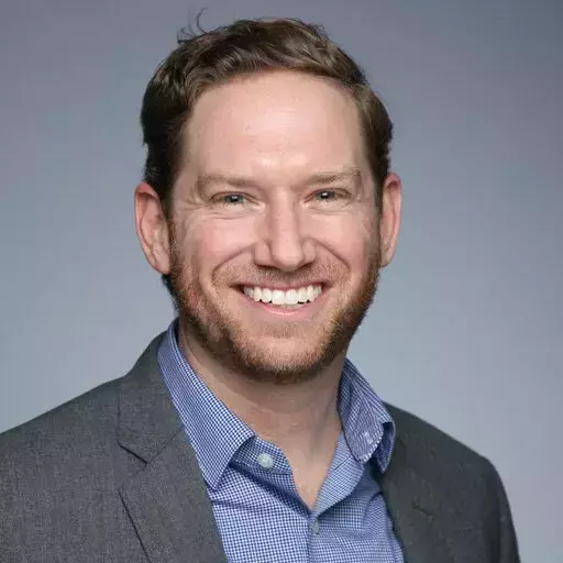 A man with reddish-brown hair and a beard smiles broadly at the camera. He wears a gray suit jacket over a blue and white checkered button-down shirt.