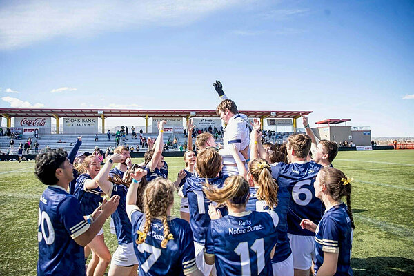 Team members in navy and gold jerseys celebrate as one player is lifted into the air.