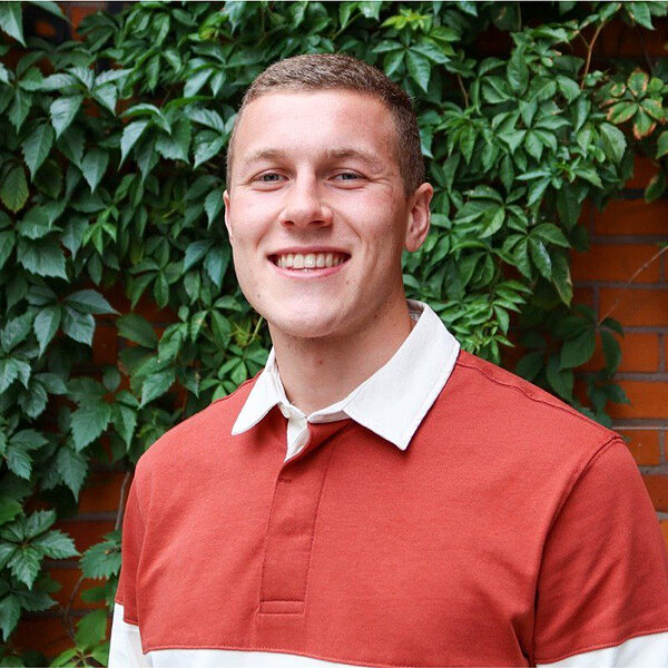 Young man with short hair smiles, wearing a rust-colored and white polo shirt.